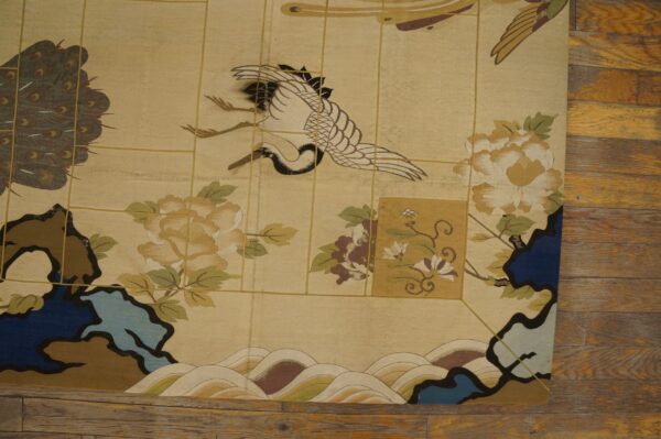 Low-pile beige rug patterned with a stylized crane, flowers, and peacock feathers on rustic wood floor.
