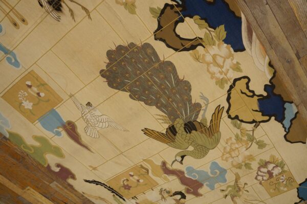 Cream rug with a detailed avian and floral design in green, brown, blue, and burgundy on distressed wood floor.