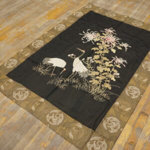 Dark textile rug features embroidered cranes and flowers, with a wide olive-gold patterned border on weathered wood flooring.