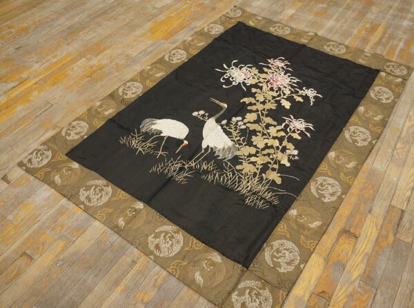 Dark textile rug features embroidered cranes and flowers, with a wide olive-gold patterned border on weathered wood flooring.