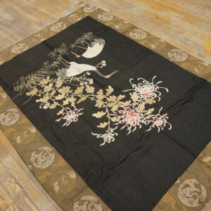 Flat black textile embroidered with white cranes and chrysanthemums, featuring a woven gold and olive border on wood flooring.