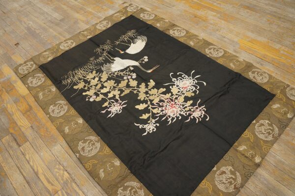 Flat black textile embroidered with white cranes and chrysanthemums, featuring a woven gold and olive border on wood flooring.