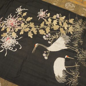 Black textile with embroidered white cranes, pink flowers, and gold leaves lies over a patterned olive rug.
