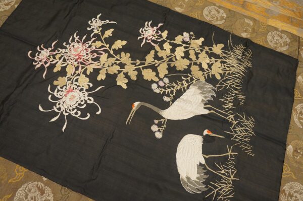 Black textile with embroidered white cranes, pink flowers, and gold leaves lies over a patterned olive rug.