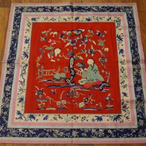 Red embroidered square textile with figures and floral motifs, framed by navy and pink patterned borders on a wood floor.
