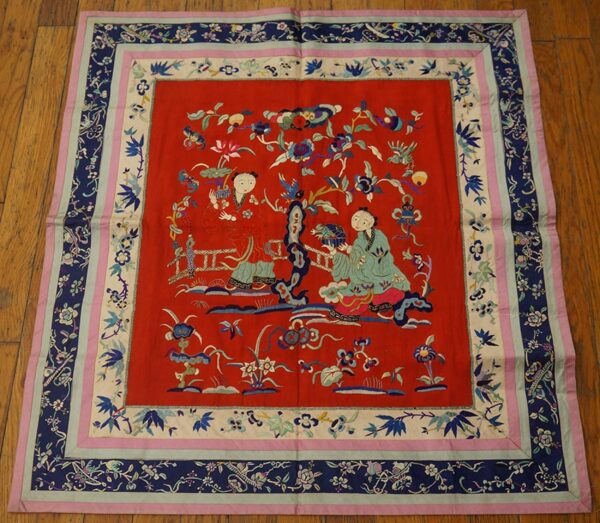 Red embroidered square textile with figures and floral motifs, framed by navy and pink patterned borders on a wood floor.