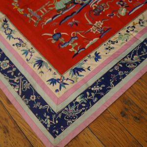 Red textile with blue, pink, and cream intricate floral border shown on distressed brown wood flooring.