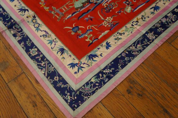 Red textile with blue, pink, and cream intricate floral border shown on distressed brown wood flooring.
