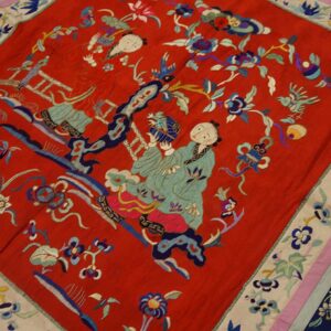 A red textile with detailed blue, teal, and pink embroidered figures and flowers, featuring a striped border.