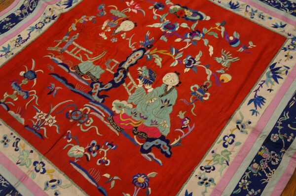 A red textile with detailed blue, teal, and pink embroidered figures and flowers, featuring a striped border.