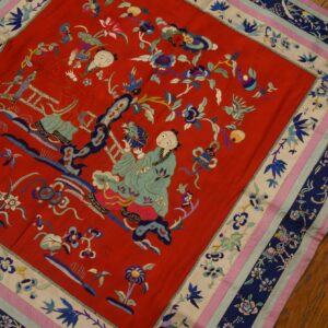 Red embroidered textile featuring two stylized figures and flowers, with a multilayered border in navy, cream, pink, and blue, set on a hardwood floor.