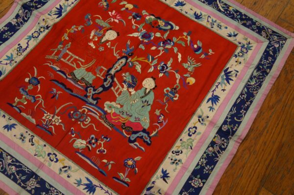 Red embroidered textile featuring two stylized figures and flowers, with a multilayered border in navy, cream, pink, and blue, set on a hardwood floor.