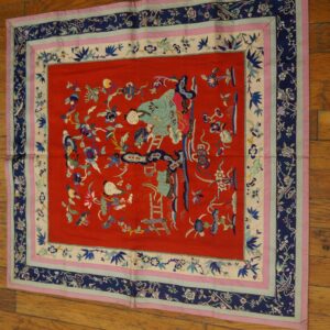 Red embroidered textile featuring figures and flowers, framed by multi-colored blue and pink layered borders on wood flooring.