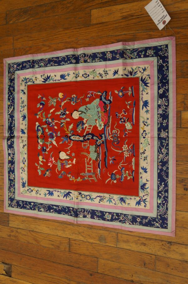 Red embroidered textile featuring figures and flowers, framed by multi-colored blue and pink layered borders on wood flooring.