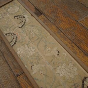 Narrow, framed textile with intricate floral and crane embroidery rests on distressed, wide plank wood flooring.