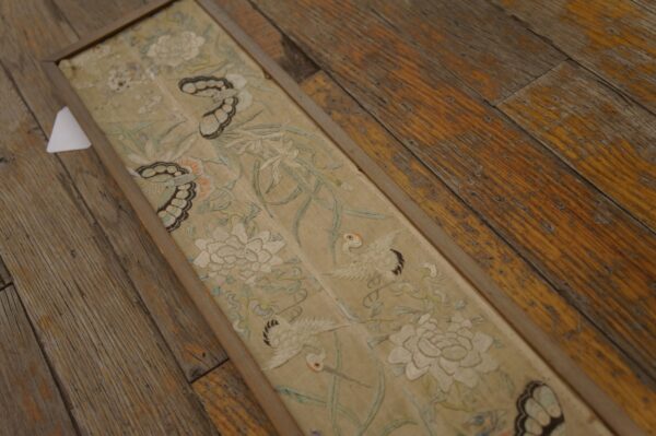 Narrow, framed textile with intricate floral and crane embroidery rests on distressed, wide plank wood flooring.