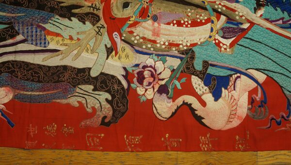 Textured textile featuring complex figures and flowers in red, teal, black, and white on a wood floor.