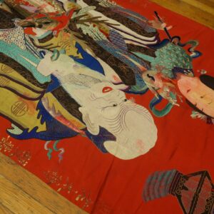 Detailed, colorful figurative textile with designs in white, gold, teal, and red resting on a light wood floor.
