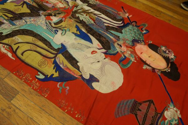 Detailed, colorful figurative textile with designs in white, gold, teal, and red resting on a light wood floor.