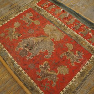 Detailed red embroidered textile with central gray lions and outer dancing figures, displayed on a wood floor.