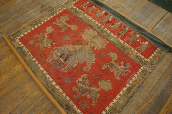 Detailed red embroidered textile with central gray lions and outer dancing figures, displayed on a wood floor.