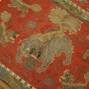 Detailed red textile featuring embroidered gray and tan mythical beasts with prominent teal eyes.
