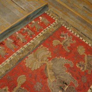 Red embroidered textile featuring detailed panels of figures and lion motifs rests on worn wood planks.
