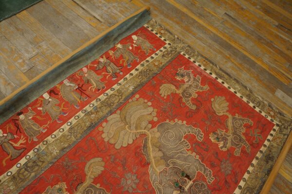Red embroidered textile featuring detailed panels of figures and lion motifs rests on worn wood planks.