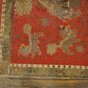 Red embroidered textile with couched gold and gray mythological figures, decorative borders, and circular mirrors on wood planks.