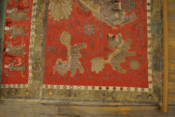 Red embroidered textile with couched gold and gray mythological figures, decorative borders, and circular mirrors on wood planks.