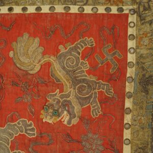 Red embroidered fabric detailed with coiled gold and gray threads showing a creature and a swastika.