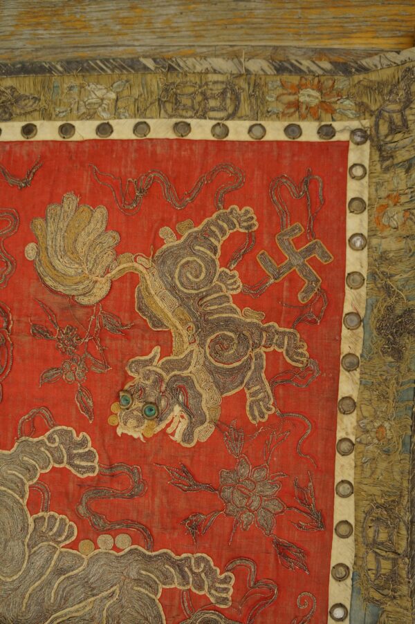 Red embroidered fabric detailed with coiled gold and gray threads showing a creature and a swastika.