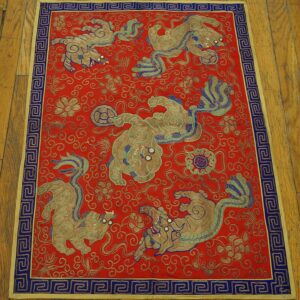Red embroidered textile featuring detailed gold animal motifs and a blue Greek key border, placed on wood floor.