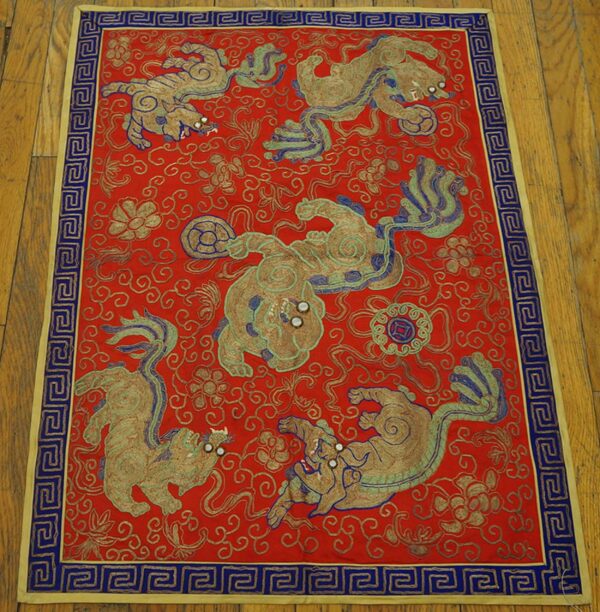Red embroidered textile featuring detailed gold animal motifs and a blue greek key border, placed on wood floor.