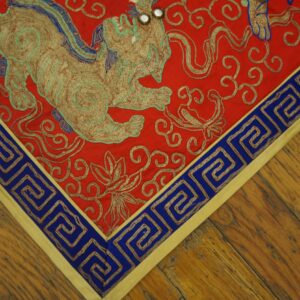Red embroidered textile featuring a gold creature, blue Greek key border, laying on a wood floor.