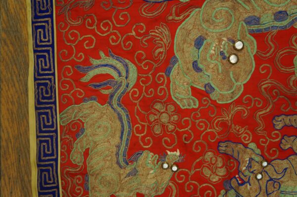 Red embroidered textile featuring gold, teal, and blue mythical figures and a meander border.
