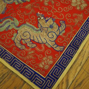 A low-pile red rug featuring a beaded gold animal motif and a blue meander border rests on wood planks.