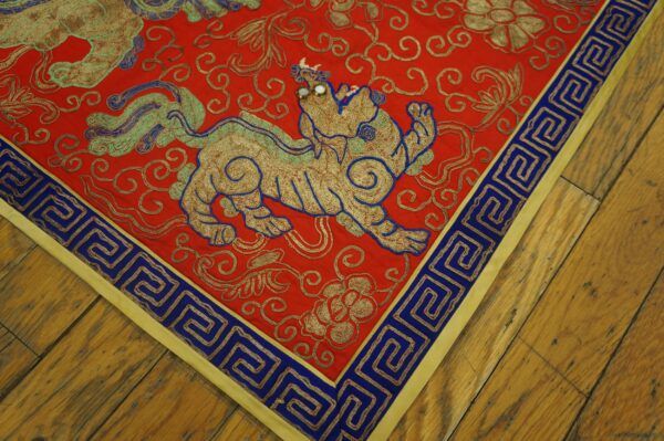 A low-pile red rug featuring a beaded gold animal motif and a blue meander border rests on wood planks.