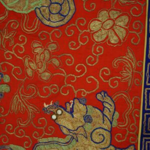 Bright red textile with gold, blue, and green embroidered motifs and a meander border lying on wood flooring.