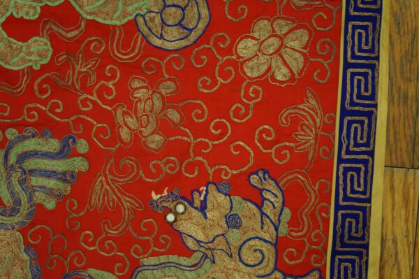 Bright red textile with gold, blue, and green embroidered motifs and a meander border lying on wood flooring.