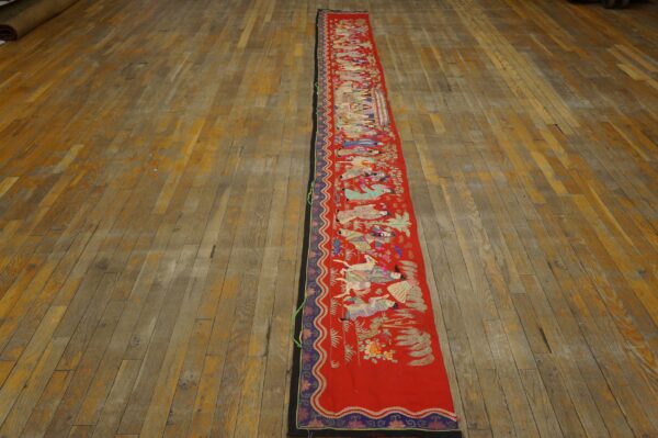Long red textile runner with colorful embroidered figures on a distressed wooden plank floor.