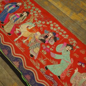 Vibrant red textile featuring densely embroidered scenes of figures and animals rests on distressed wood flooring.