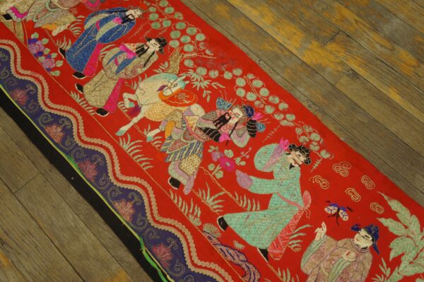 Vibrant red textile featuring densely embroidered scenes of figures and animals rests on distressed wood flooring.