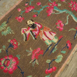 Brown textile runner with embroidered pink floral and figure patterns on distressed wooden flooring.