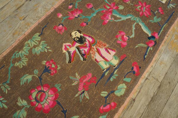 Brown textile runner with embroidered pink floral and figure patterns on distressed wooden flooring.