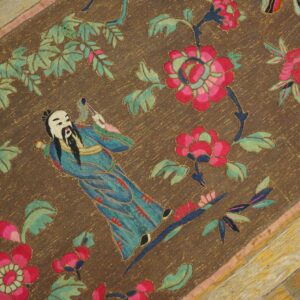 Detailed flat weave brown rug featuring a blue figure, pink flowers, and green leaves on weathered wood flooring.