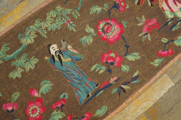 Detailed flat weave brown rug featuring a blue figure, pink flowers, and green leaves on weathered wood flooring.