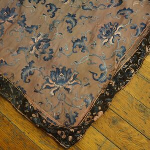 Corner view of a tan textile with blue floral embroidery and a dark patterned border on wood floor.
