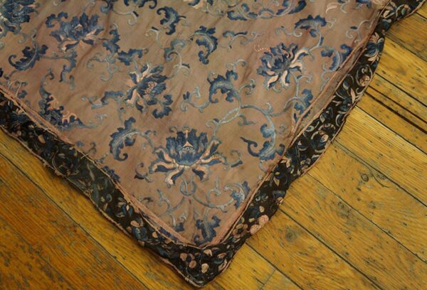 Corner view of a tan textile with blue floral embroidery and a dark patterned border on wood floor.