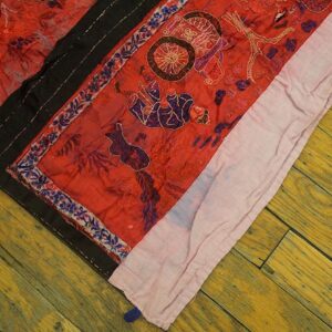 Crinkled red textile with dark purple and gold stitched patterns, black borders, lying on distressed yellow wood floor.